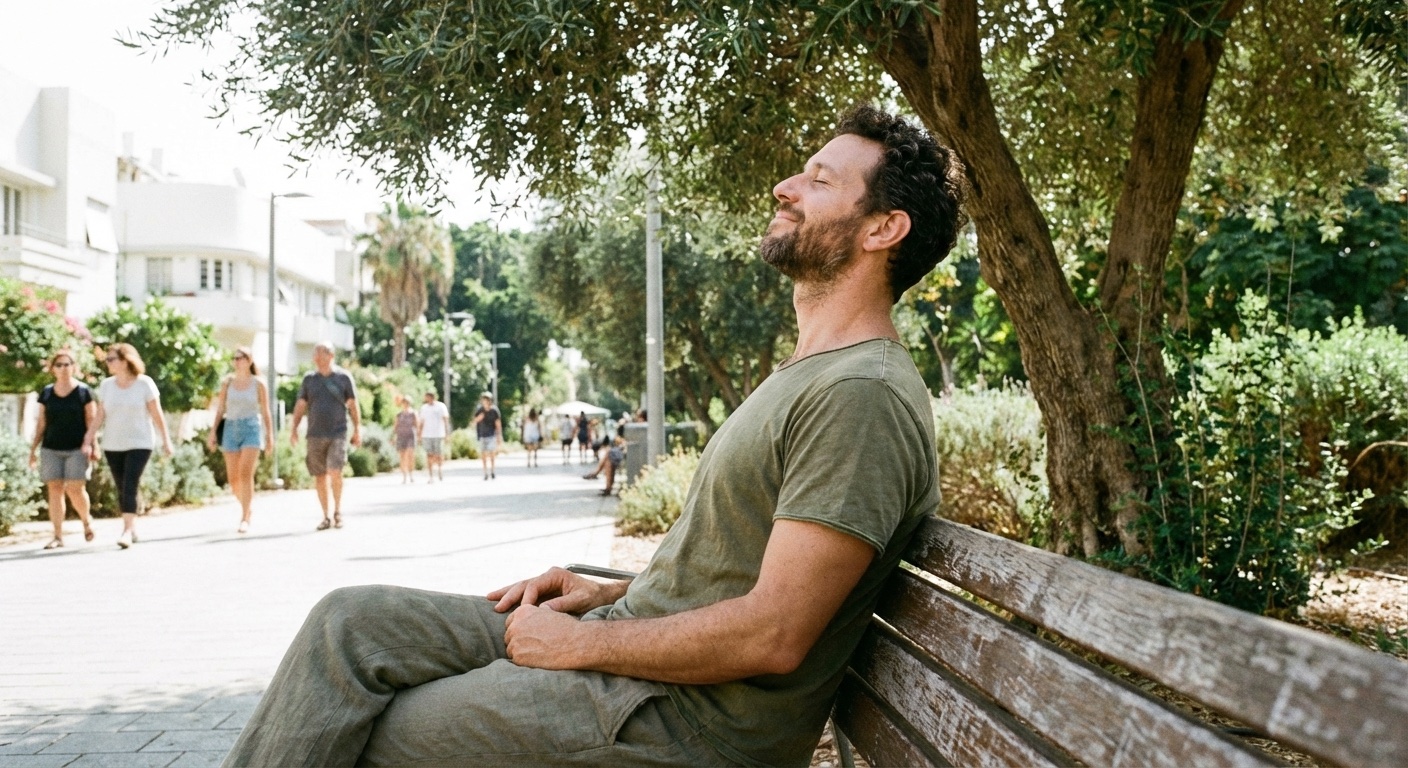 גבר ישראלי צעיר יושב על ספסל בפארק עם עיניים עצומות, נושם עמוק ונראה רגוע